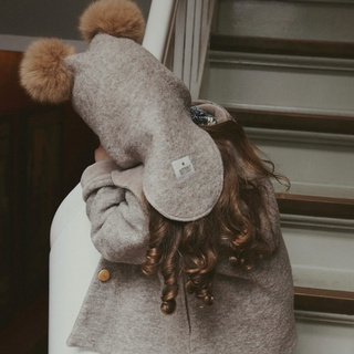 A child wearing a light brown wool coat and a matching brown hat with two pom-poms, leaning against a white chair in front of wooden stairs.