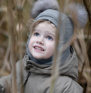 A young child wearing a gray knit hood with large pom-poms and a taupe winter jacket, peeking through dry reeds outdoors.