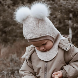 A toddler wearing a beige winter coat and a cream knit pom-pom hat with a neck warmer, looking down while holding a wicker basket outdoors.