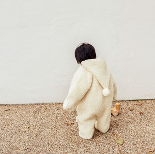 Child in a snugglesuit with a hood, standing on a sandy surface.