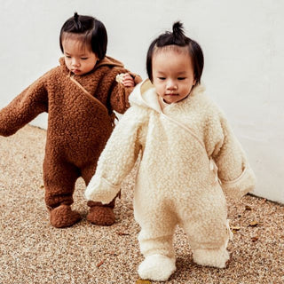 Two children in brown and beige snugglesuit onesies standing on a textured surface.