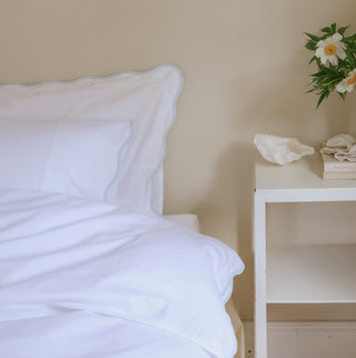 White bedding with blue trim detail on a bed with a nightstand holding decorative items in a bedroom setting.