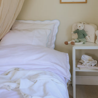 Bedroom with a bed with white bedding with blue trim detail  and nightstand featuring a teddy bear and books.