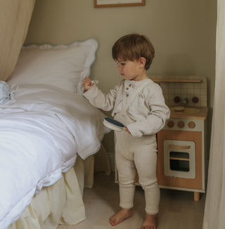 Child in pajamas standing near a bed with white bedding and blue trim detail with a toy kitchen set in the background