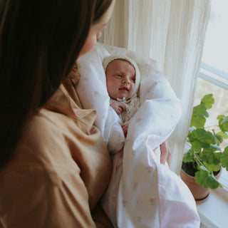 Woman holding a baby wrapped in a white blanket with pink florals by a window with plants.