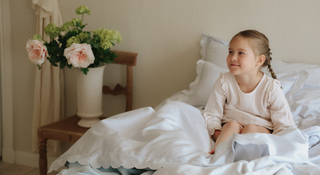 Young girl sitting on a bed with a vase of flowers on a side table.