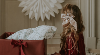 Child with a large white bow in hair standing next to a gift box with a floral patterned pillow.
