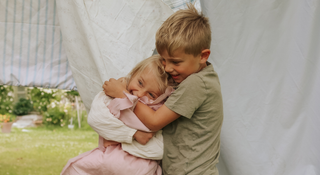 Two children hugging in a garden setting with a white tent in the background.