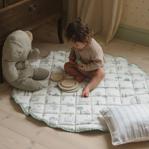 Adventures in Wonderland playmat in Aqua. Child playing on top of playmat with pillow, plates and stuffed animal