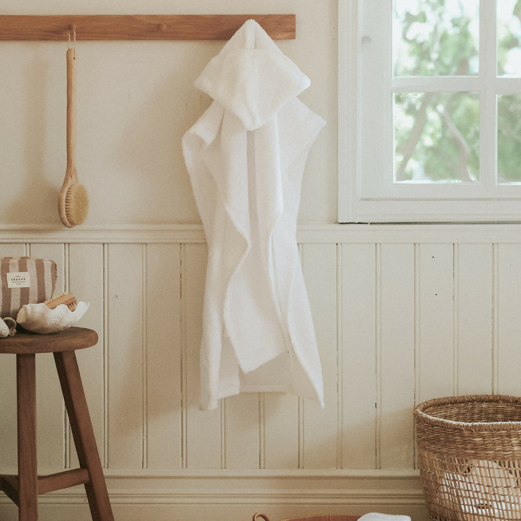 Toddler Poncho hanging up on the wall in a bathroom. A wooden stool with bath accessories to the left and a rattan storage basket with towels to the right.