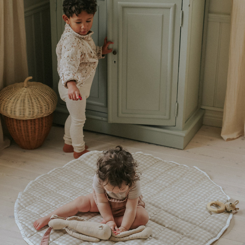 Picnic Gingham Playmat in Beige. Child sitting on playmat with stuffed animal. Other child is standing behind her watching her play