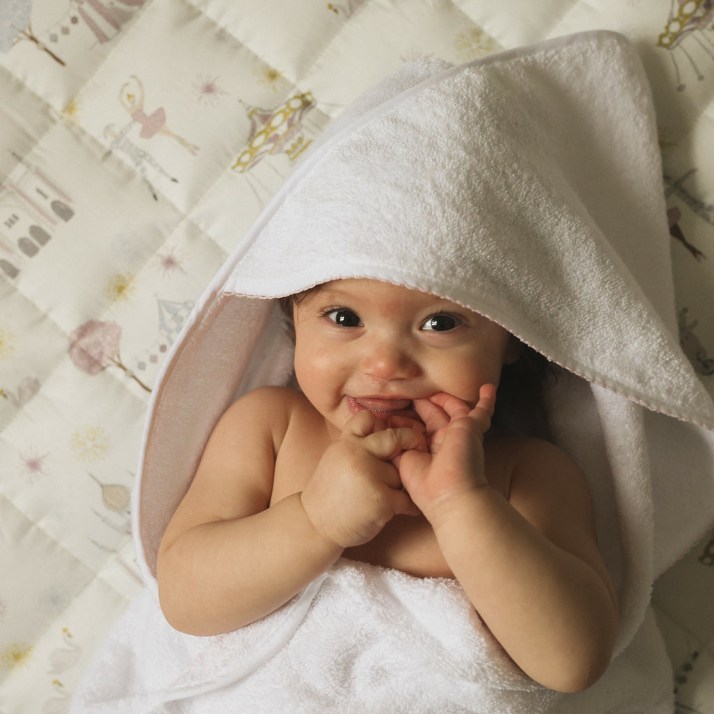 Baby Hooded Towel in white with pink trim detail. Baby laying on playmat smiling