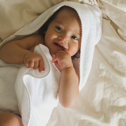 Baby hooded towel in white with beige trim. Baby is  wrapped in hooded towel while laying on bed smiling