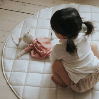 Child sitting on a round, quilted mat with a plush toy on a wooden floor.