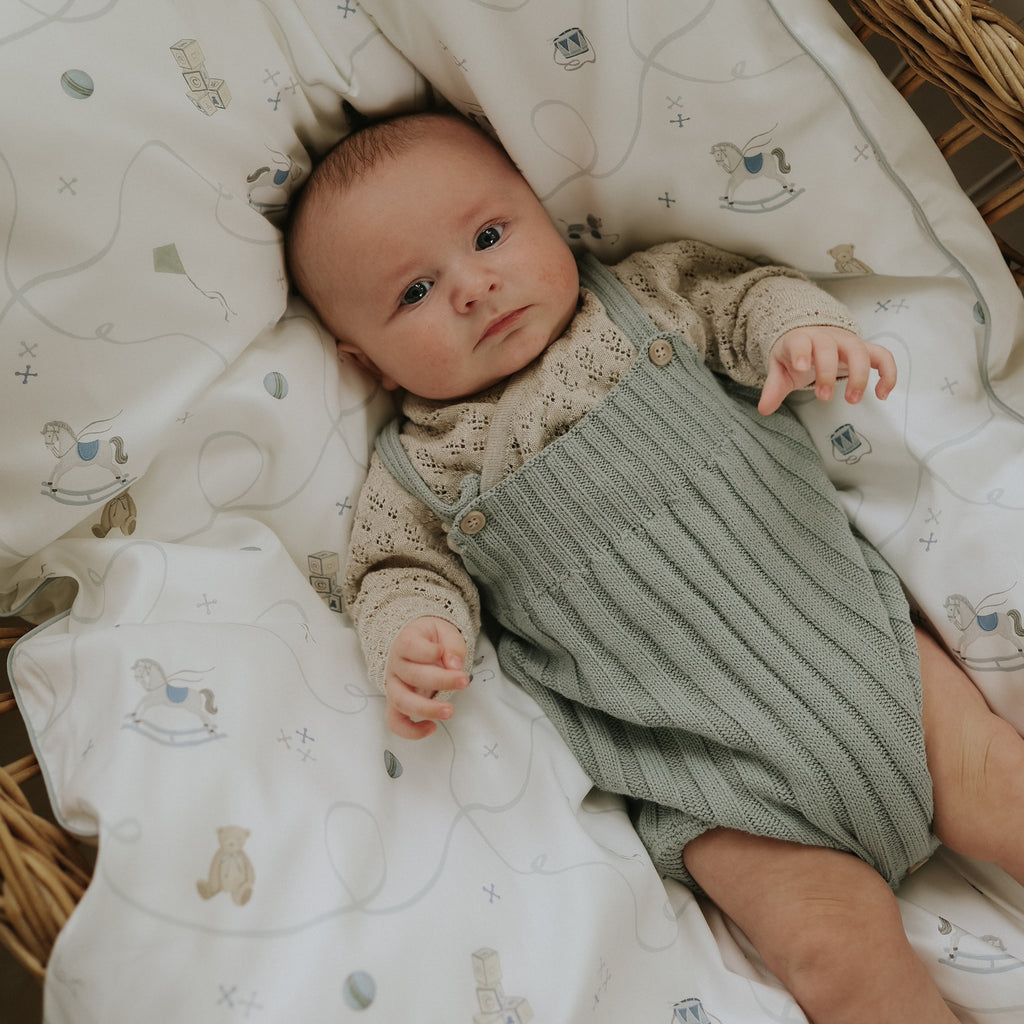 Baby lying on a patterned blanket wearing a green romper with a beige shirt.
