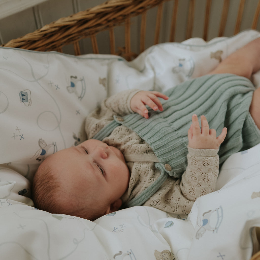 Baby lying in a crib wearing a green and beige outfit with a soft blanket.