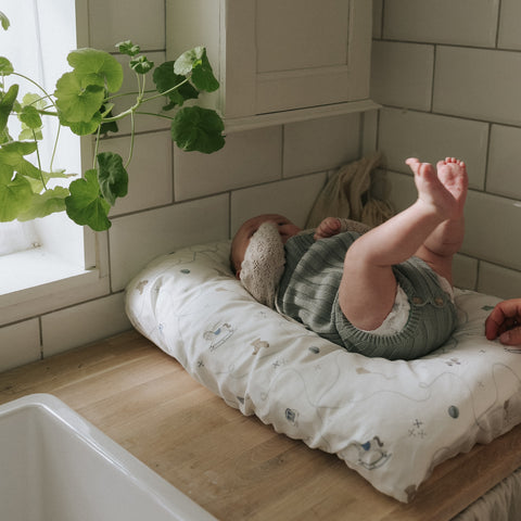 Baby lying on a changing pad in a kitchen with a plant and tiled wall in the background