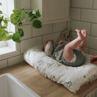 Baby lying on a changing pad in a kitchen with a plant and tiled wall in the background