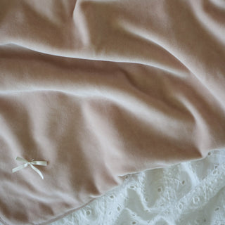 A close-up of a soft blush pink velvet throw blanket with a small white bow, draped over a white lace sheet.