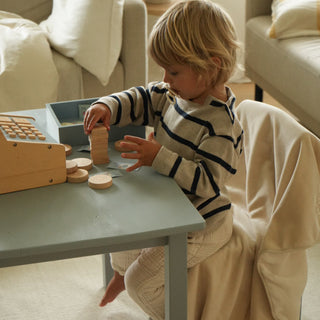 A young child sits on a cream upholstered chair at a small light blue table, playing with wooden toy blocks and a toy cash register in a softly lit living room.