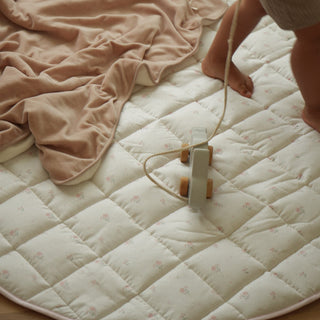A baby's feet and legs are visible as they pull a small wooden toy cart across a large, cream-colored diamond-quilted play mat with a dusty pink blanket on a light wood floor.