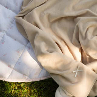A beige fleece blanket with a small white embroidered cross detail, draped over a light blue floral quilted blanket on green grass.
