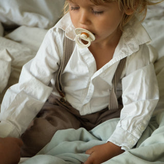 A young child wearing a white long-sleeve shirt, brown suspenders, and brown pants sits on a bed with a pale blue blanket, holding a pacifier to their mouth.