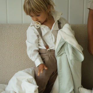 A toddler wearing a white shirt and brown suspenders sits on a bed, wrapped in a pale sage green blanket. The scene is styled with neutral, soft-toned bedding and a textured beige headboard.