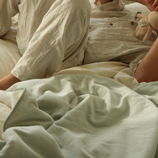 A person reclining on a bed wearing loose, light beige pajamas and a matching top, with rumpled pale green bedding in the foreground and a softly lit bedroom background.