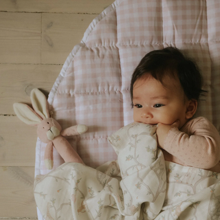 Baby lying on a pink gingham playmat with a plush toy, surrounded by soft fabrics.