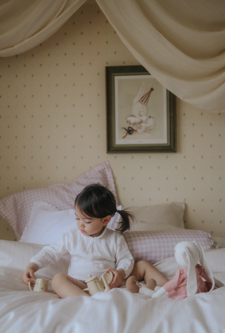 Child sitting on a bed with toys, wearing a white outfit, in a room with decorative wallpaper and a framed picture.