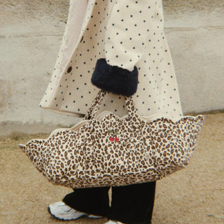 A young child wearing a cream polka dot coat with dark navy trim, a matching beanie, and holding a leopard print doll bassinet, standing outdoors in front of a stone wall.