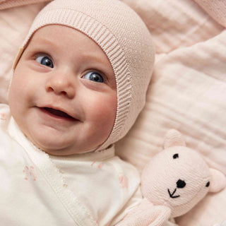 A close-up of a smiling baby wearing a pale pink knit bonnet and a cream bodysuit, lying on a soft pink blanket and holding a plush bear toy.