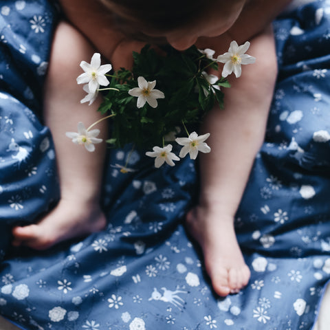 Toddler sitting on the gooselings duvet in the print "Into The Woodlands" in the color blue, holding flowers in hand